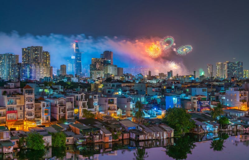 Colorful fireworks light up the night sky above a city skyline with tall buildings, as smoke drifts over the scene. In the foreground, houses and buildings reflect in a calm river.
