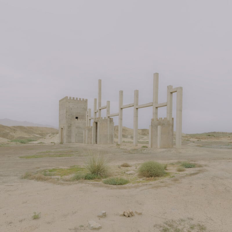 A partially constructed or abandoned concrete structure with tall pillars and a castle-like tower stands in a barren, desert landscape under a cloudy sky. Sparse vegetation grows on the sandy ground.
