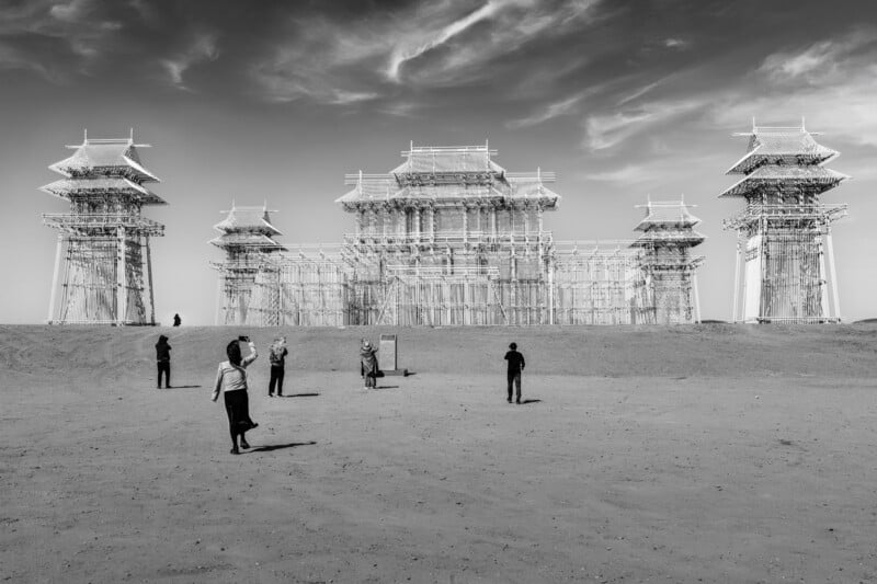 Black and white photo of people standing in front of a large, ornate structure made of scaffolding, resembling a historic building with towers, set in a barren, sandy landscape under a partly cloudy sky.