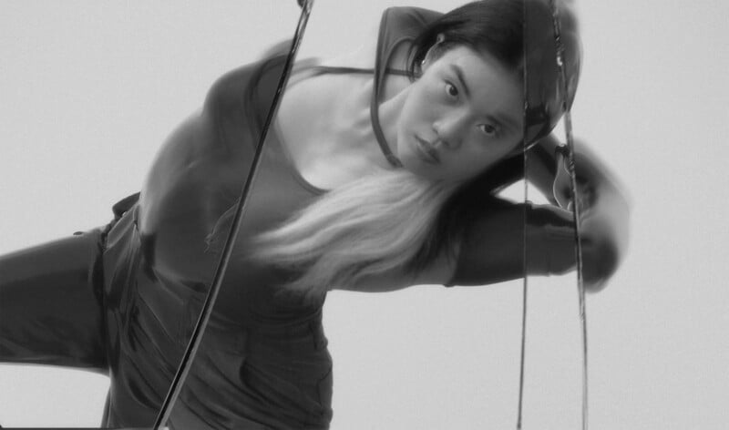 A person with long, partially blonde hair poses in a dynamic, leaning position. They wear a fitted, dark outfit and look directly at the camera. The image is black and white and has a blurred, distorted effect.