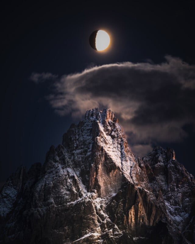 A dramatic mountain peak with snow is illuminated by moonlight under a dark sky. The bright moon, partially obscured by shadow, appears above the mountain through scattered dark clouds.