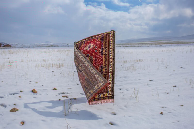 A colorful, patterned rug stands upright and folded in a snowy, open field with mountains and a cloudy sky in the background. Sparse dry grass pokes through the snow.
