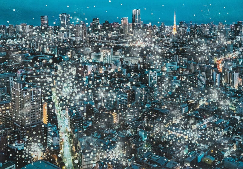 A vibrant cityscape at night with numerous tall buildings and skyscrapers illuminated by lights. White dots are scattered across the image, giving a dreamy, sparkling effect. The sky is a deep blue.