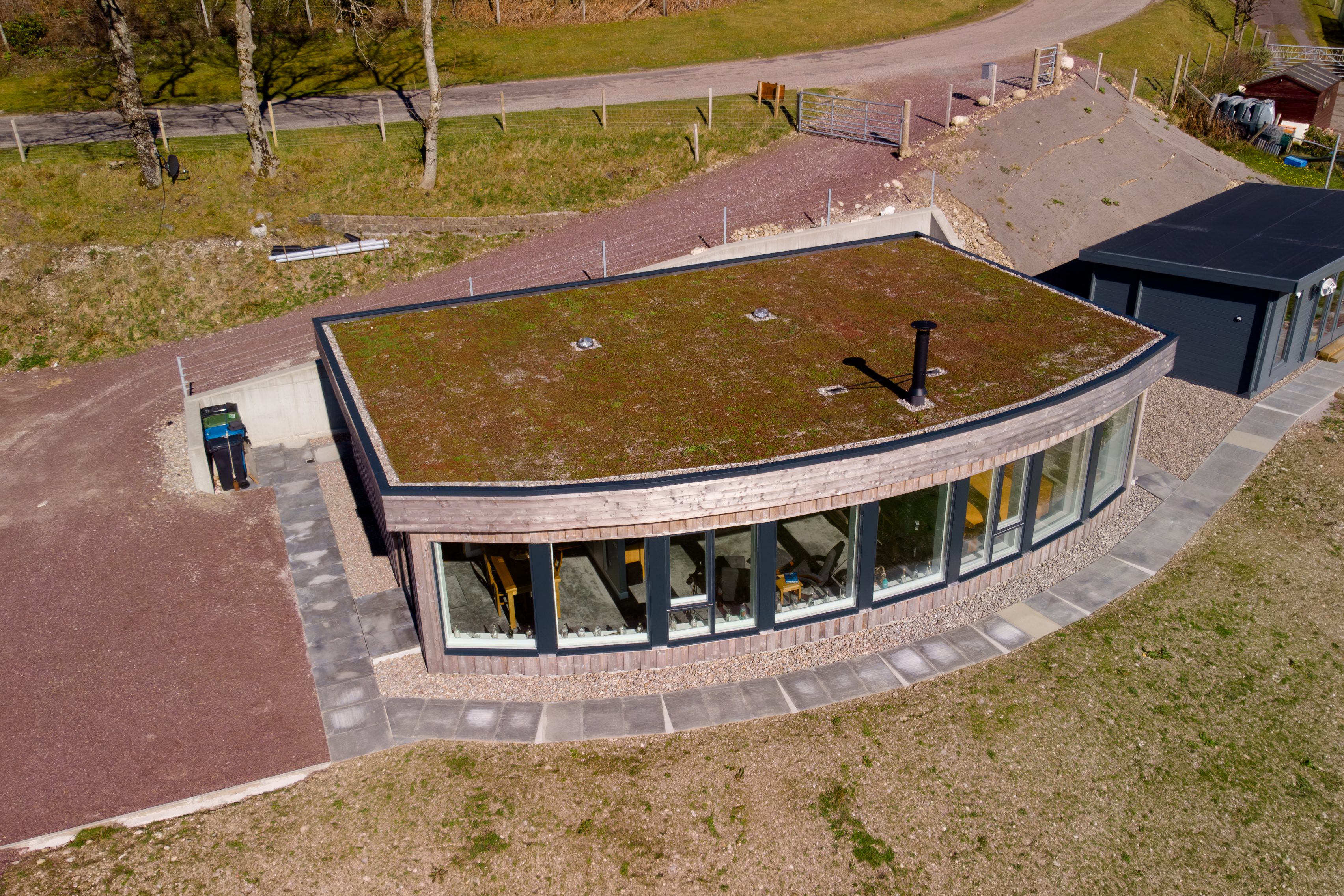 Aerial view of a contemporary building with a green roof, curved wooden facade, and large windows, surrounded by gravel paths and dry grass.