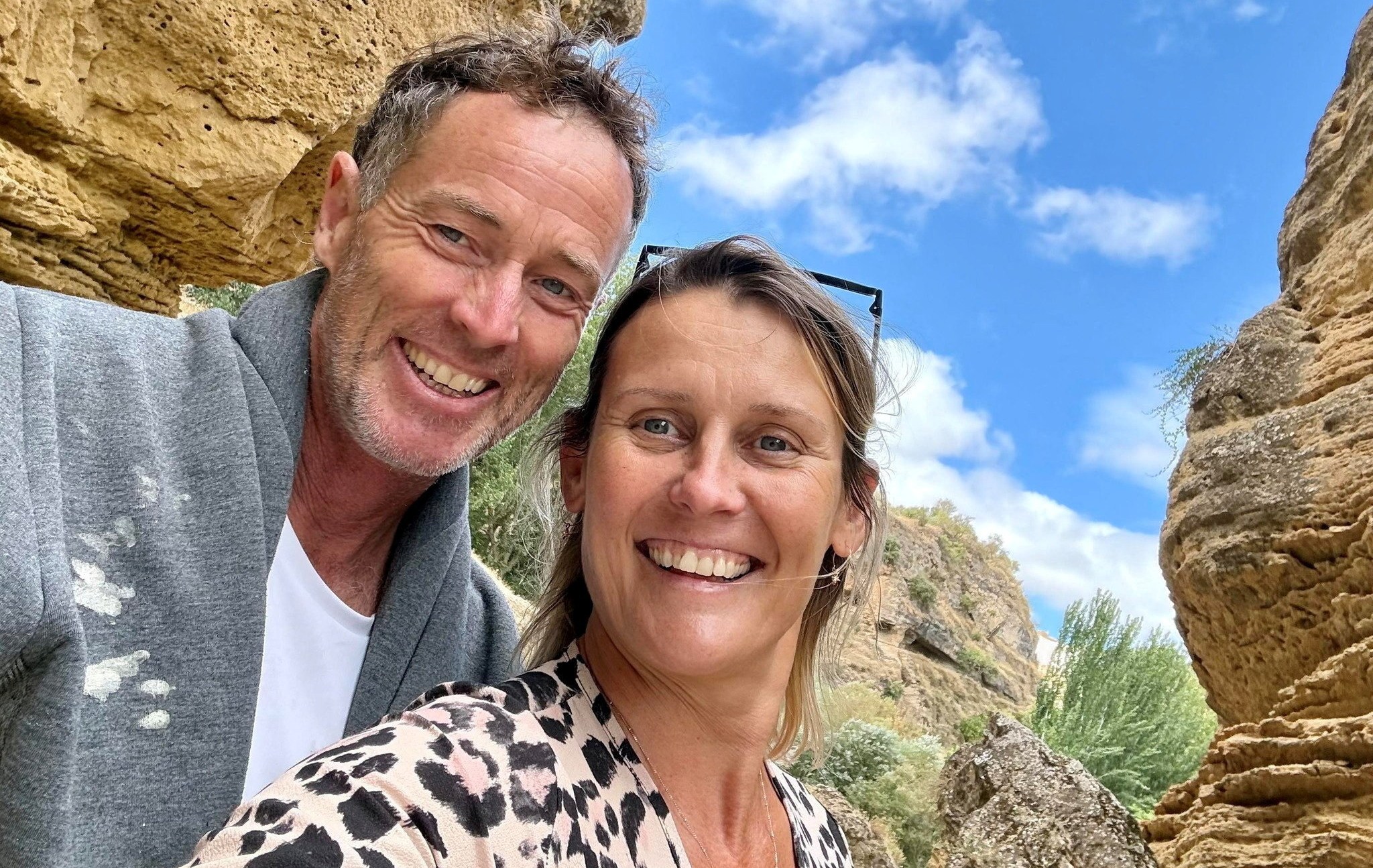 Craig and Lindsay Foreman pose for a selfie outdoors with rock formations and a blue sky behind them.
