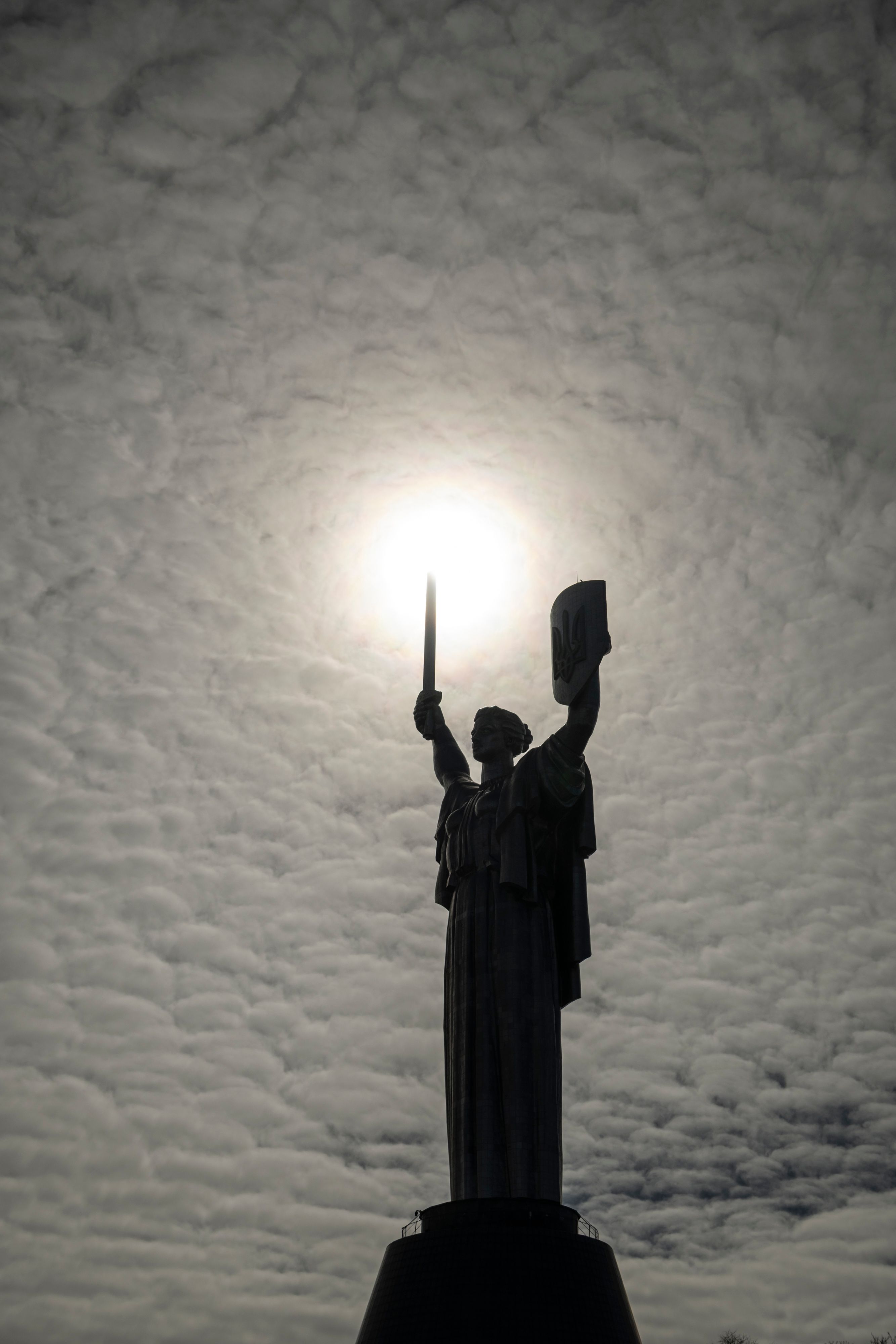 The Mother of Ukraine statue, a female figure holding a sword and shield, silhouetted against a cloudy sky with the sun shining brightly behind it.