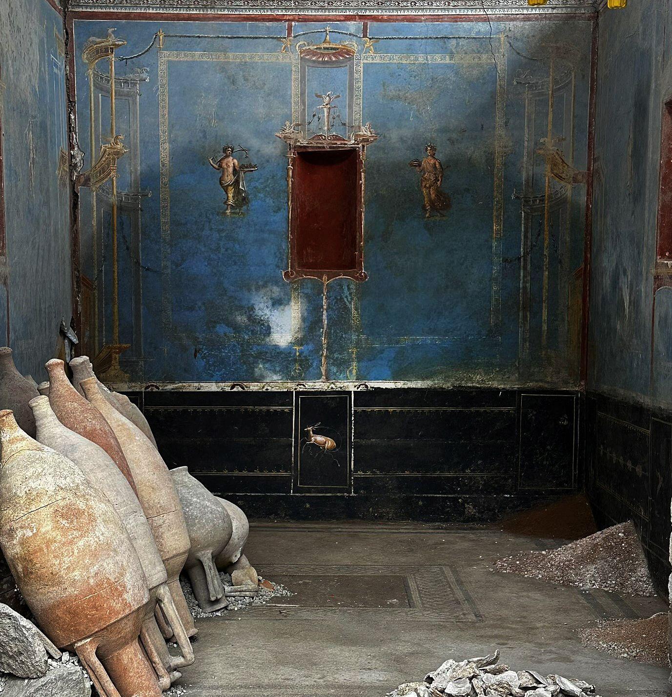Excavated Pompeii sacrarium with blue frescoed walls, several large amphorae, and piles of debris.