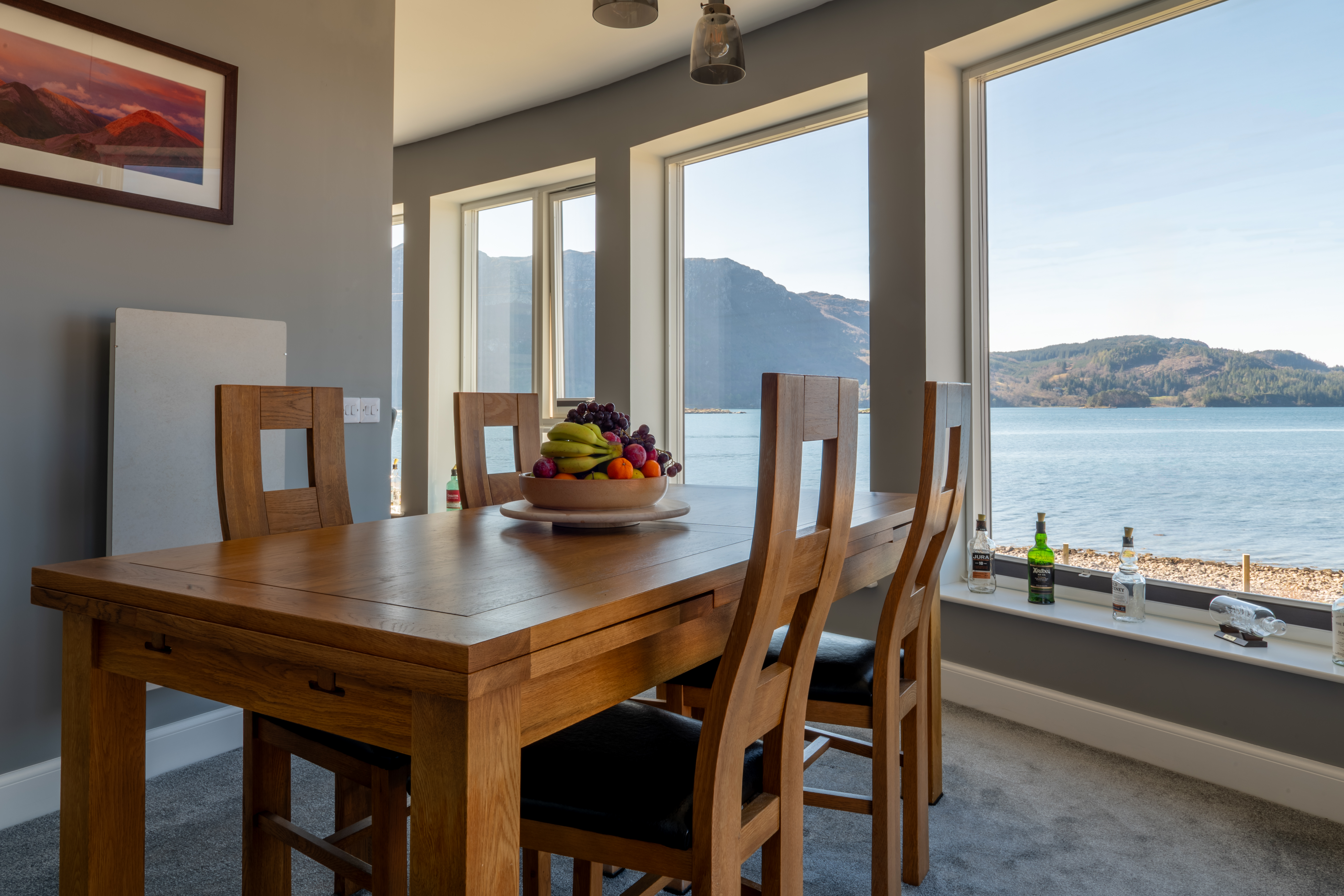 A wooden dining table with chairs and a fruit bowl in a room with large windows overlooking a loch with mountains and a forested shoreline.