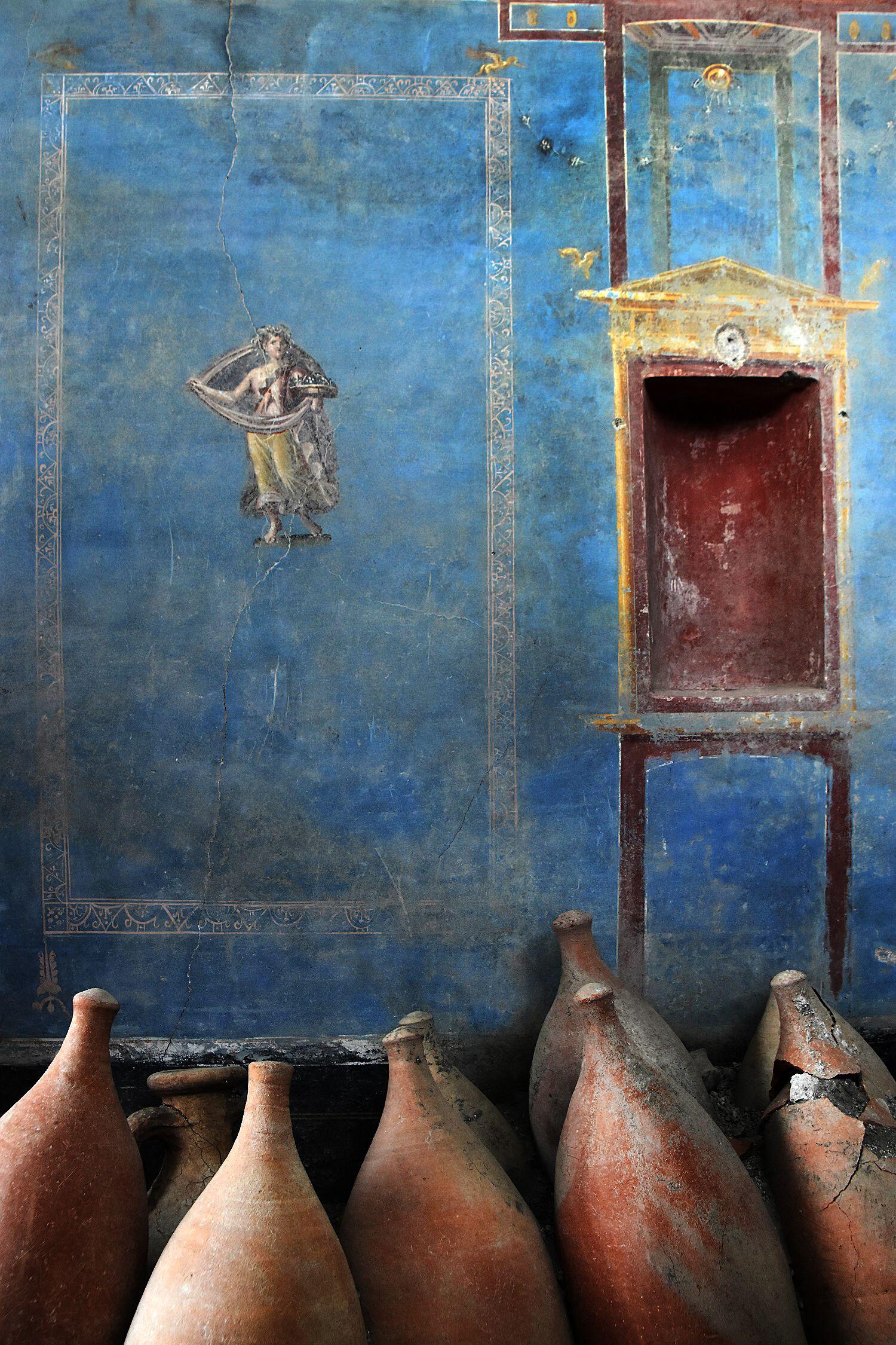 Ancient blue fresco with a female figure and decorative niche, with several clay amphorae in the foreground.