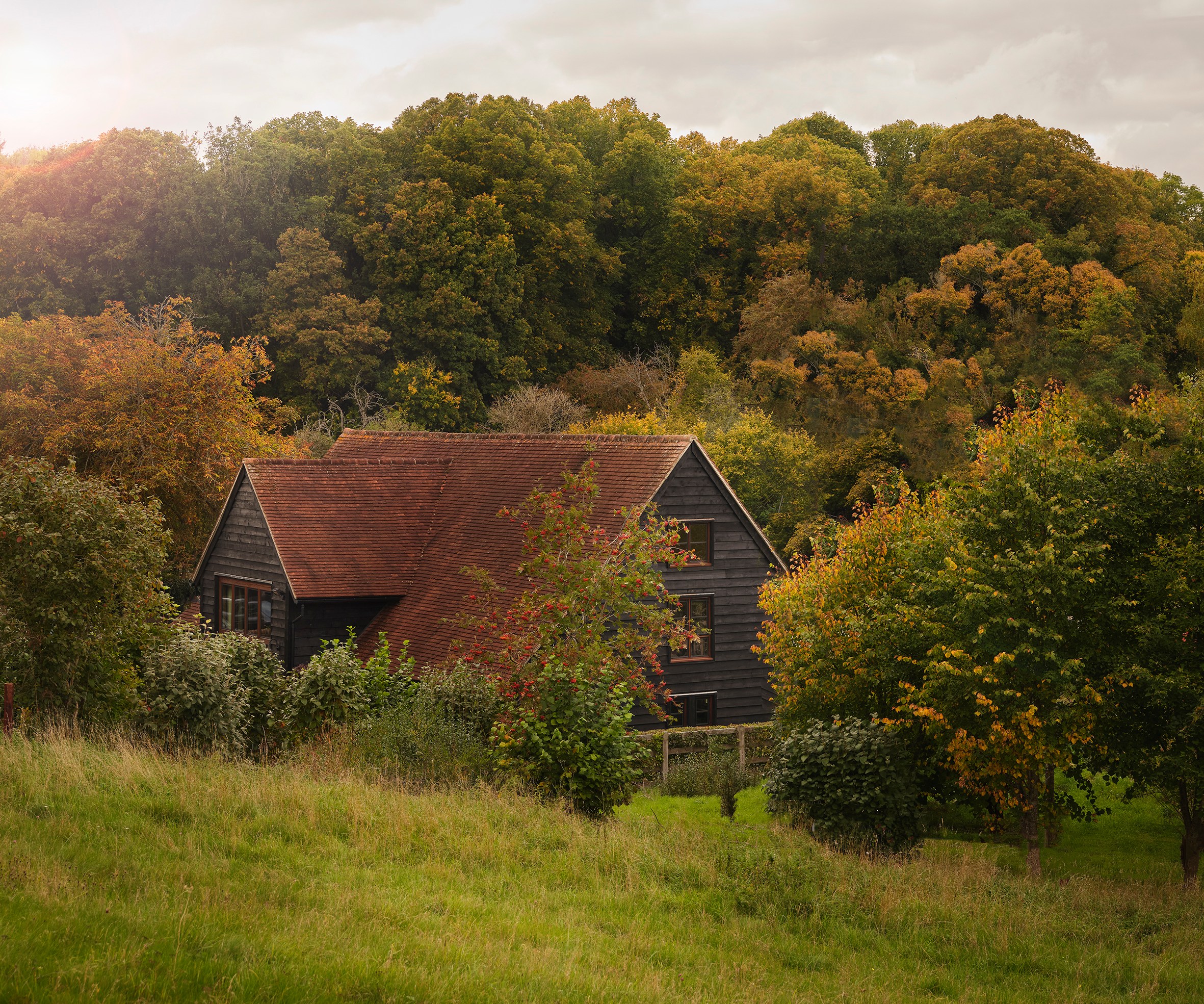 A dark wooden house with a red-tiled roof nestled among trees with green and yellow foliage, under a cloudy sky.