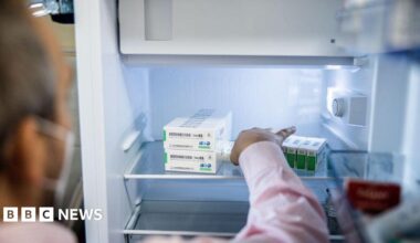 A woman with dark hair and a multiple colour jumper siting on a green sofa inside her living room.