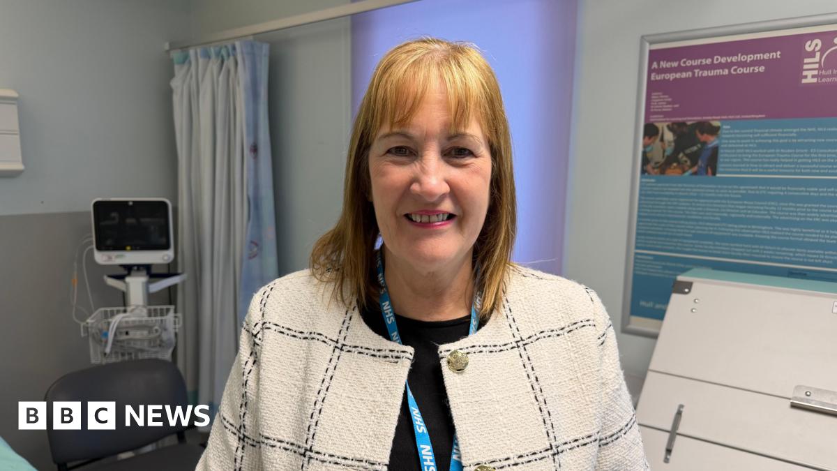A woman wearing a light-coloured, checked jacket and an NHS lanyard stands in a hospital room. Medical equipment, a curtained area and information boards are visible in the background.