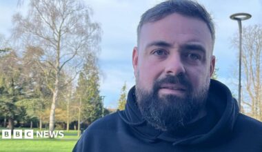 A man with black hair and beard who is wearing a black hooded top. There is a park behind him with trees and grass and someone is walking a dog.