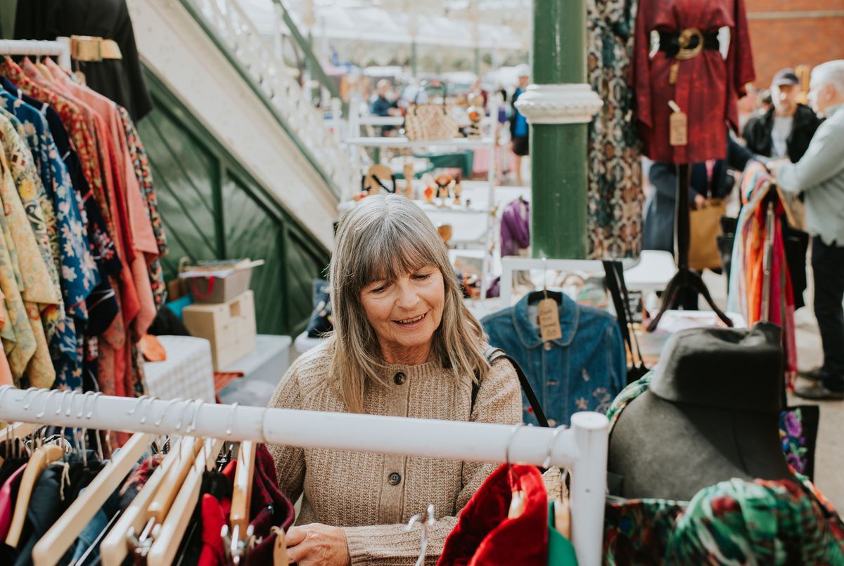 A glamorous older woman happily shops at a clothing stall in an indoor market on a sunny day.