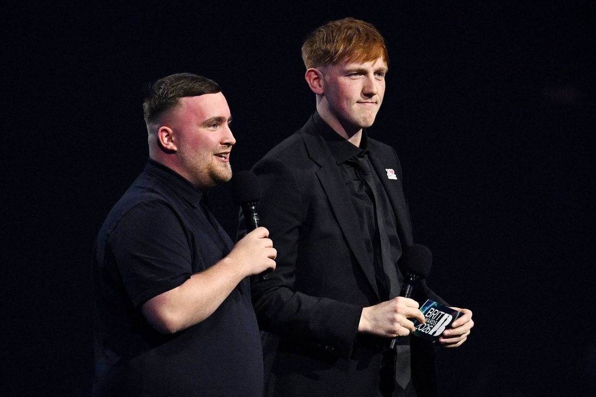 Two individuals are standing on a stage, likely at an awards ceremony. One person is holding a microphone and appears to be speaking, while the other individual is holding an award and attentively listening. The background is dark, creating a spotlight effect on the two individuals.