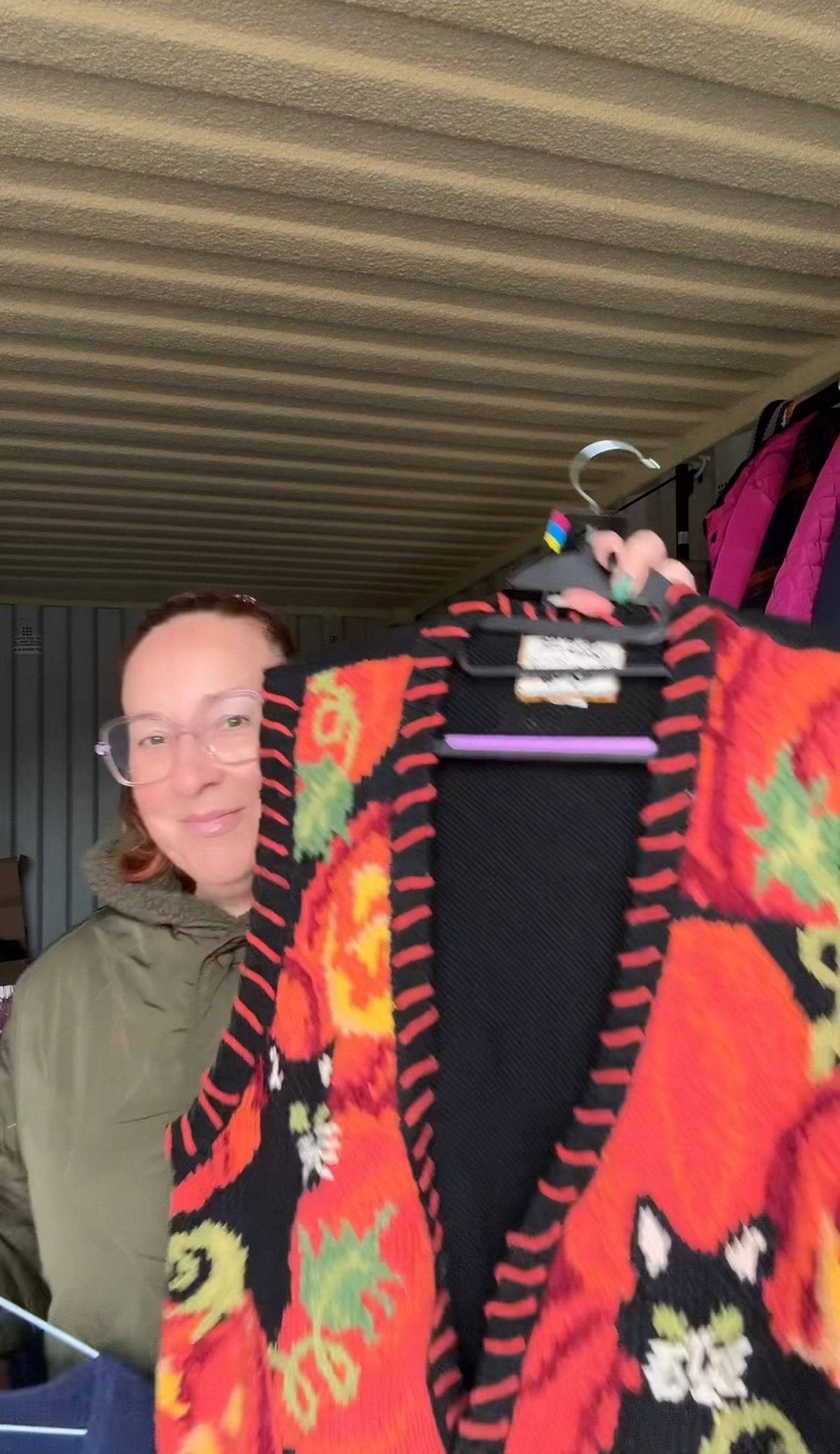 An individual wearing glasses and a green jacket is standing in a room with a metal ceiling. They are holding up a vibrant red and black garment featuring a black cat design and various autumnal motifs, which is hanging on a hanger.