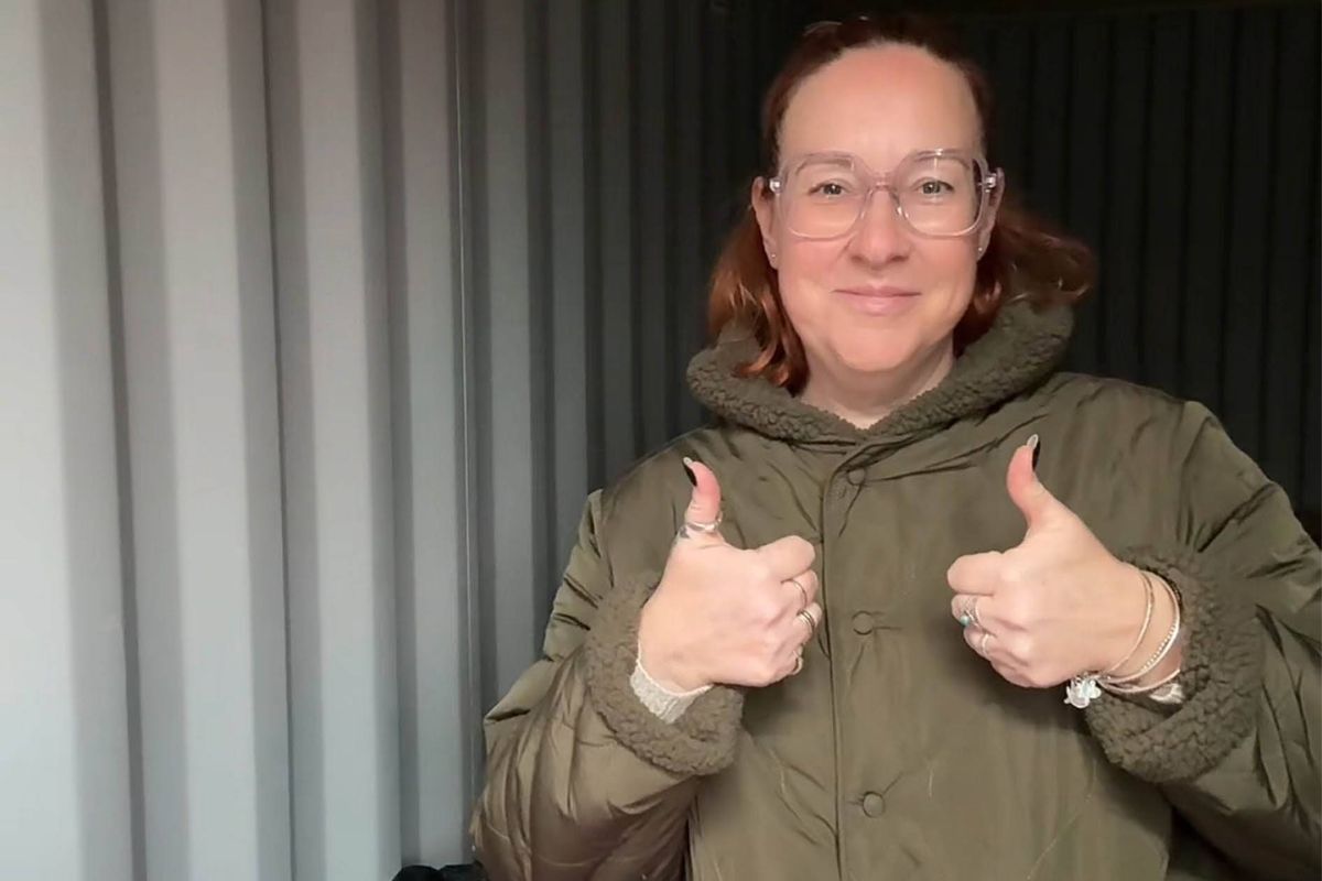 An individual dressed in a green jacket with a hood is standing and giving a thumbs-up gesture with both hands, positioned in front of a wall with vertical stripes.