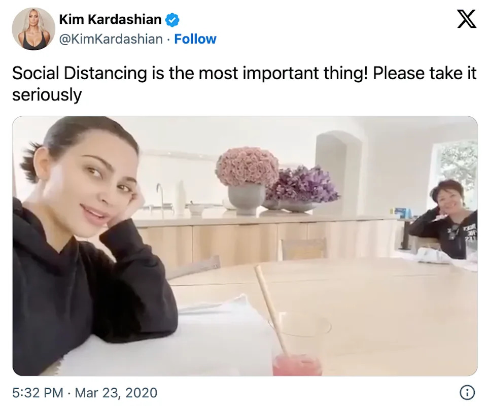 Kim Kardashian shares a social media post about social distancing, sitting at a table with a family member in a modern kitchen