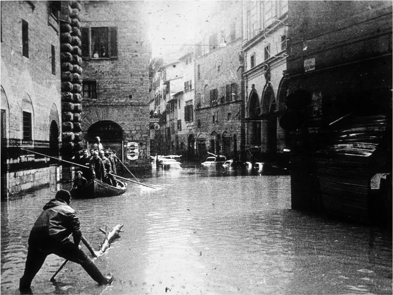 A man in the foreground uses a large stick to move through the floodwaters, while a boat full of people passes in the background during the 1966 Florence flood.