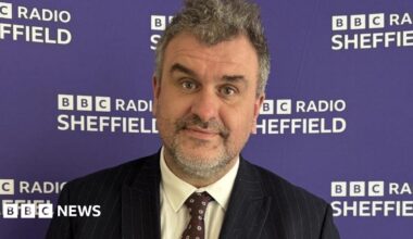 A middle-aged man, with grey hair and beard, wears a pin-stripe black suit with a brown tie. He is standing against a purple BBC Radio Sheffield backdrop.