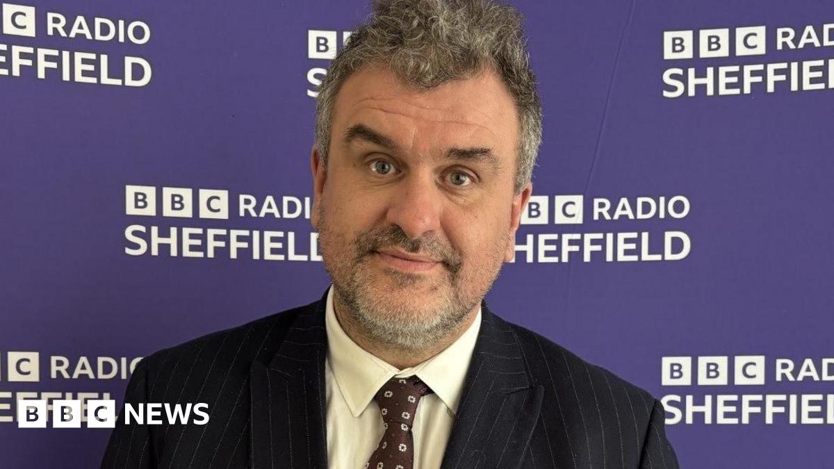 A middle-aged man, with grey hair and beard, wears a pin-stripe black suit with a brown tie. He is standing against a purple BBC Radio Sheffield backdrop.