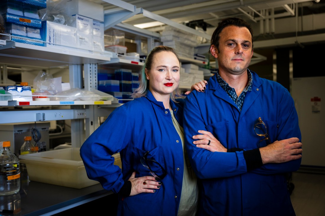 Abigail Koppes and Ryan Koppes pose for a portrait together in blue labcoats, standing in front of shelves of lab equipment.