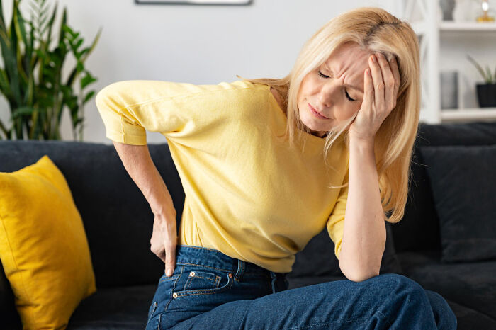 Woman in yellow top sitting on couch holding her back and head, depicting patients faking it with pain symptoms.