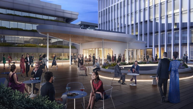 People socialize and dine on a modern outdoor terrace at dusk, surrounded by sleek, multi-story office buildings with large windows and lush planters. Some guests sit at tables, while others stand and chat near the central building.