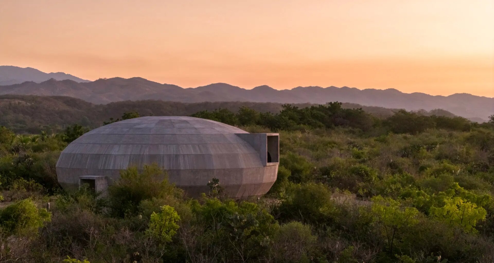 Concrete dome-shaped house in lush green landscape at sunset with mountains in the background