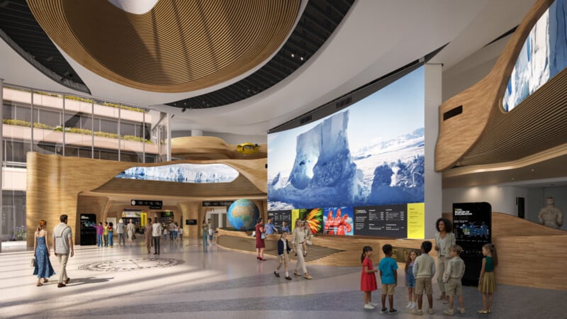 A spacious, modern museum lobby with large curved wooden structures, digital screens displaying nature scenes, a globe display, and visitors, including families and children, exploring the interactive exhibits.