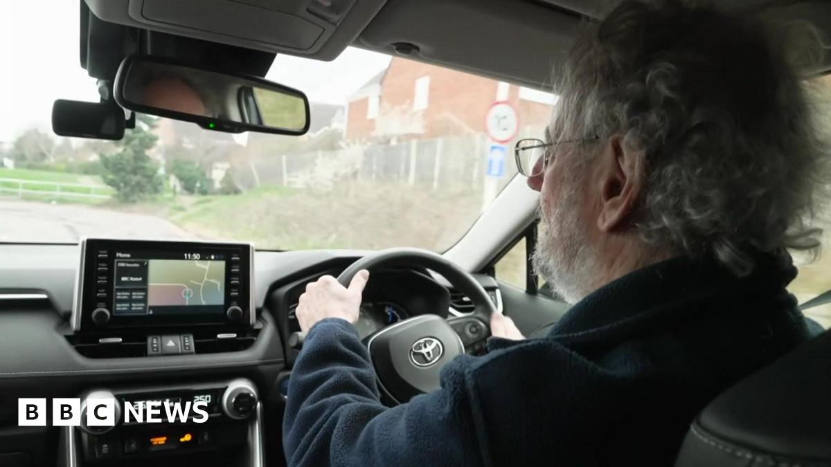 Photograph of Turner driving, taken over his shoulder. You can see the back of Turner's head - he has short, curly grey hair and a beard. He is wearing a pair of thin-framed glasses, and a dark blue fleece. The interior of his car is black, with a silver Toyota logo on the wheel. Outside the windscreen, you can see Turner is driving down a residential street.