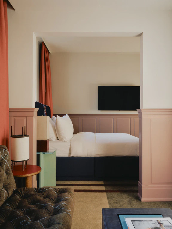 Cozy hotel room with a plush bed, wall-mounted TV, and colorful decor accents including a green side table and red curtains.