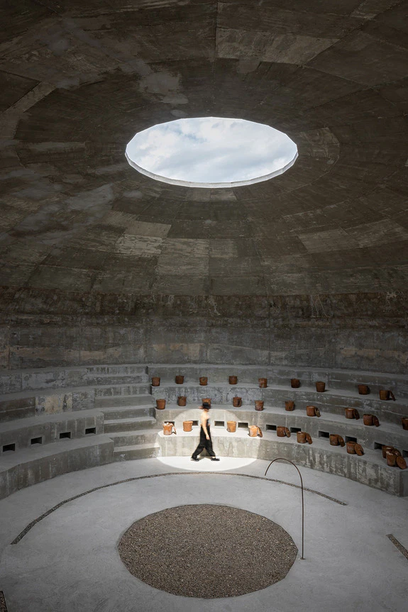 Person walking in a modern circular concrete amphitheater with a large skylight above.