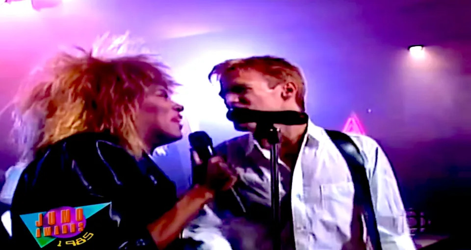 Two musicians perform energetically on stage, one with spiky hair holding a mic, the other in a white shirt playing guitar. Logo indicates 1985