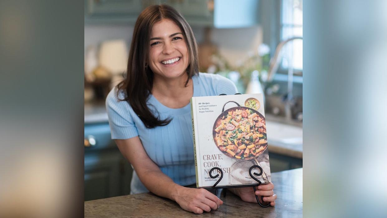 Registered dietitian Steph Grasso smiling in kitchen holding her new cookbook, "Crave, Cook, Nourish."