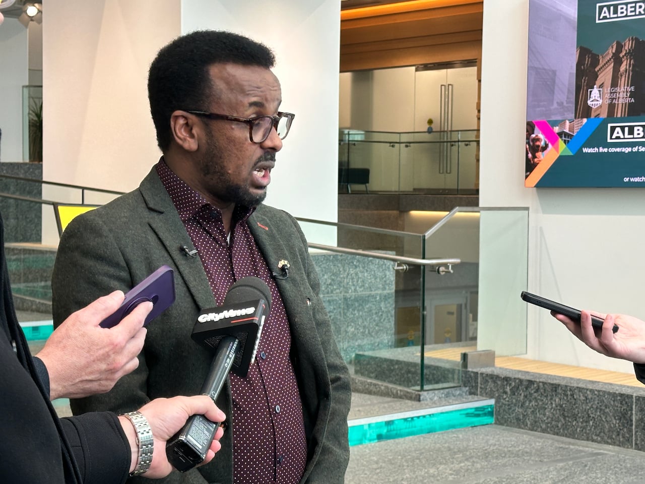 A man with black hair and glasses, wearing a maroon shirt and grey blazer, spears into audio recorders and microphones.