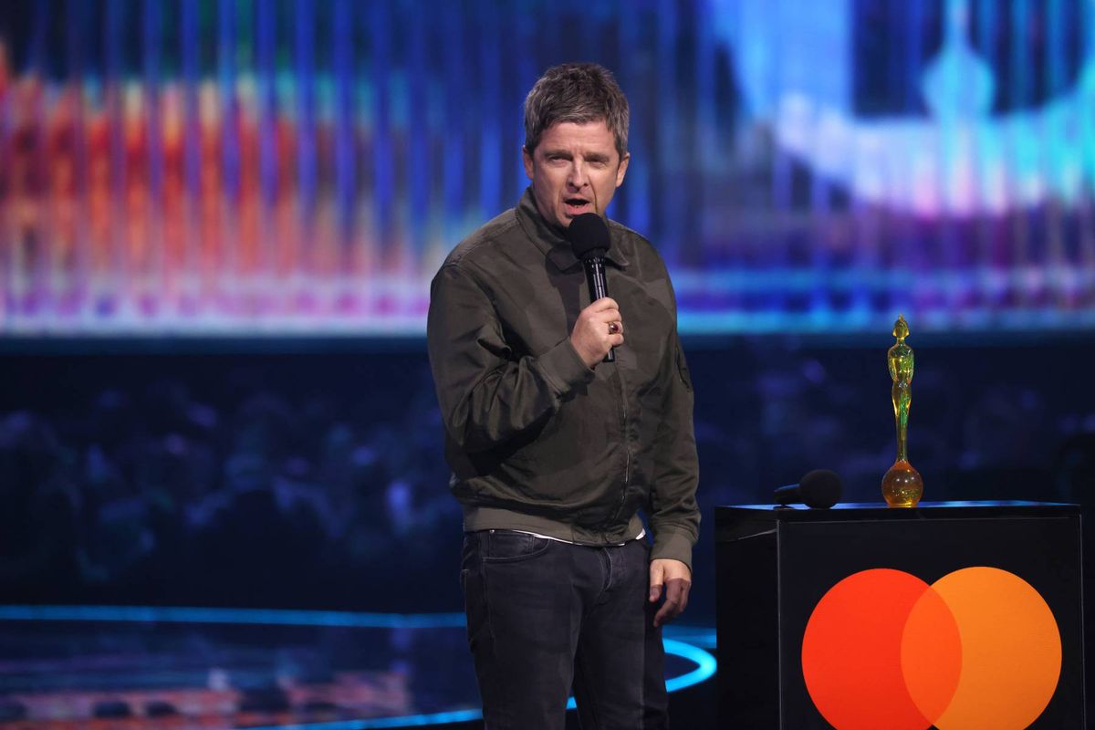 An individual is standing on a stage, holding a microphone, and appears to be delivering a speech or presentation. The stage is illuminated with blue and white lighting.