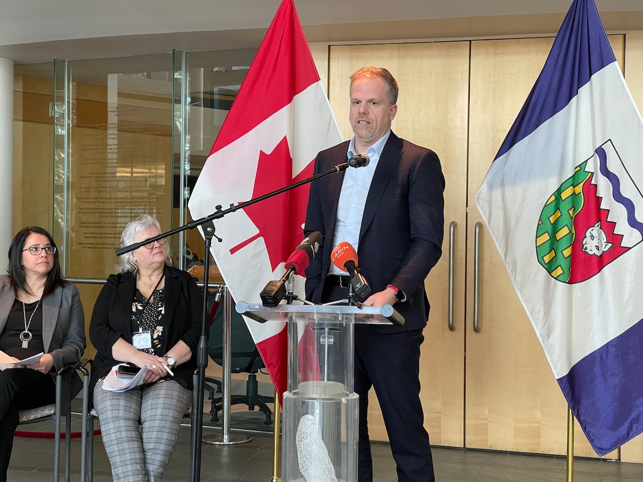 A man stands at a podium speaking into a microphone, in front of a Canadian flag and an N.W.T. flag, as two people seated beside him look on.
