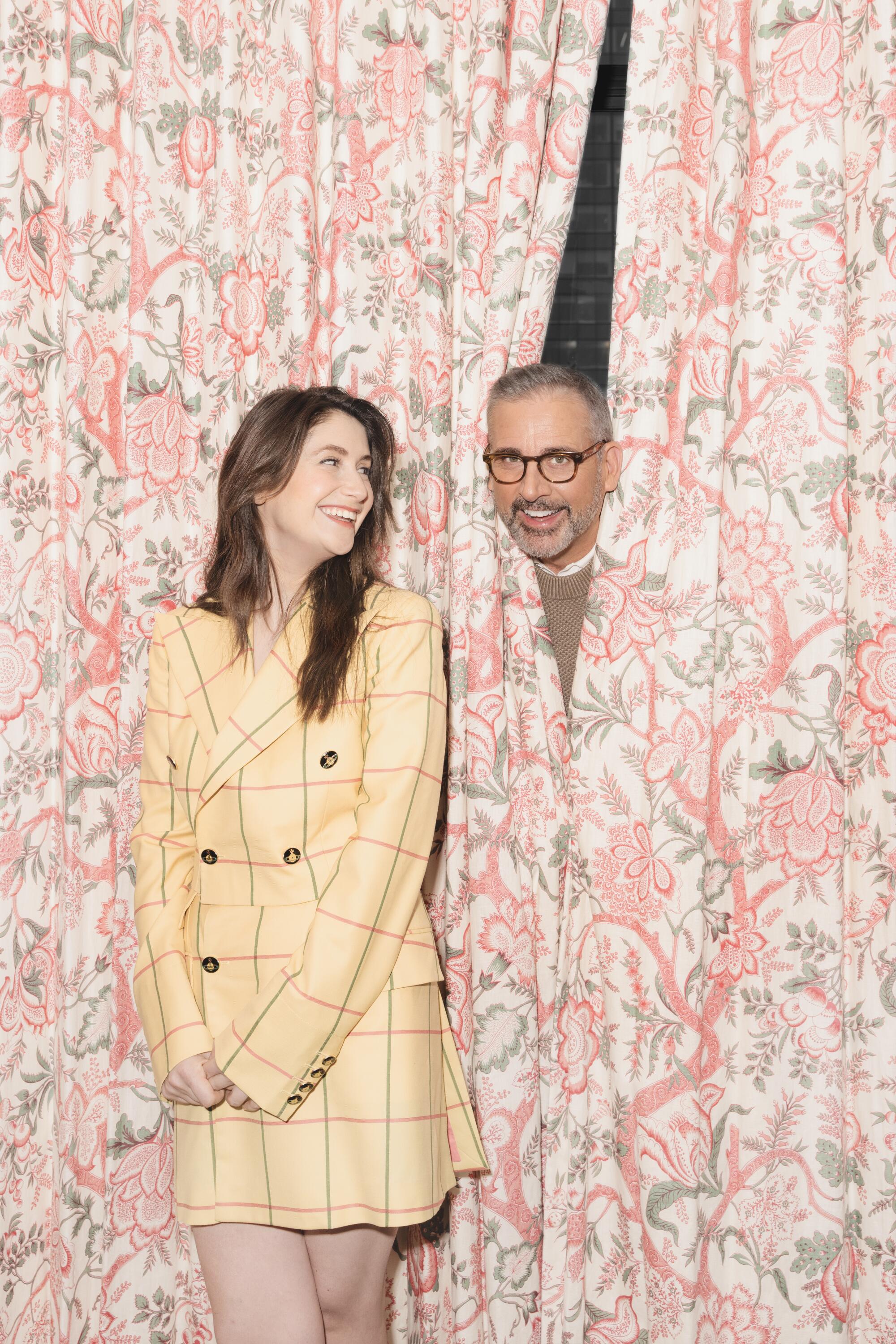 A woman in a yellow dress smiles at a man peeking out of brightly patterned drapes.