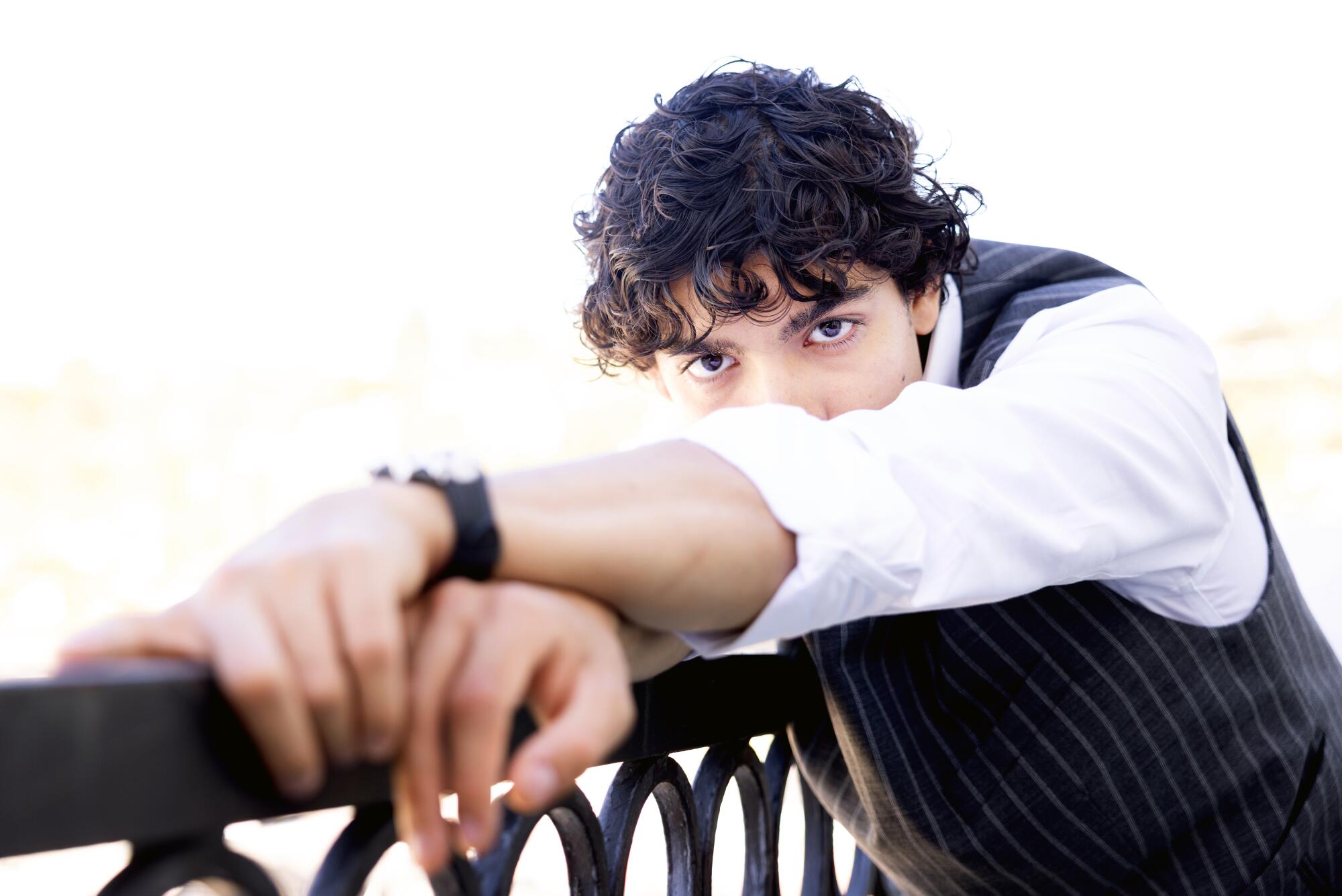 A man with curly hair leans against a black railing, his face partial covered by his arms.