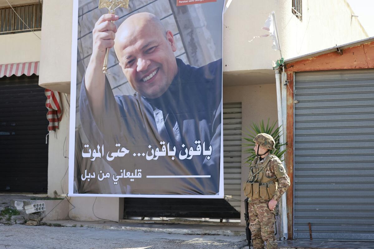 A soldier in uniform stands next to a large poster of a smiling man with a clean-shaven head, one hand raised