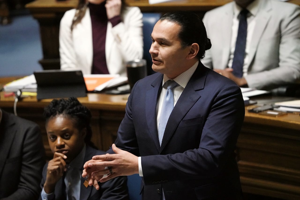 A man wearing a dark blue suit, with a dark hair in a ponytail, speaks during a government legislative session.