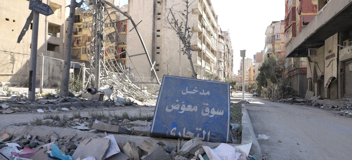 A street scene in Dahiyeh, Beirut, Lebanon, showing extensive destruction from air strikes. Buildings are heavily damaged and covered in rubble and debris. A blue sign with Arabic text lies amidst the wreckage.