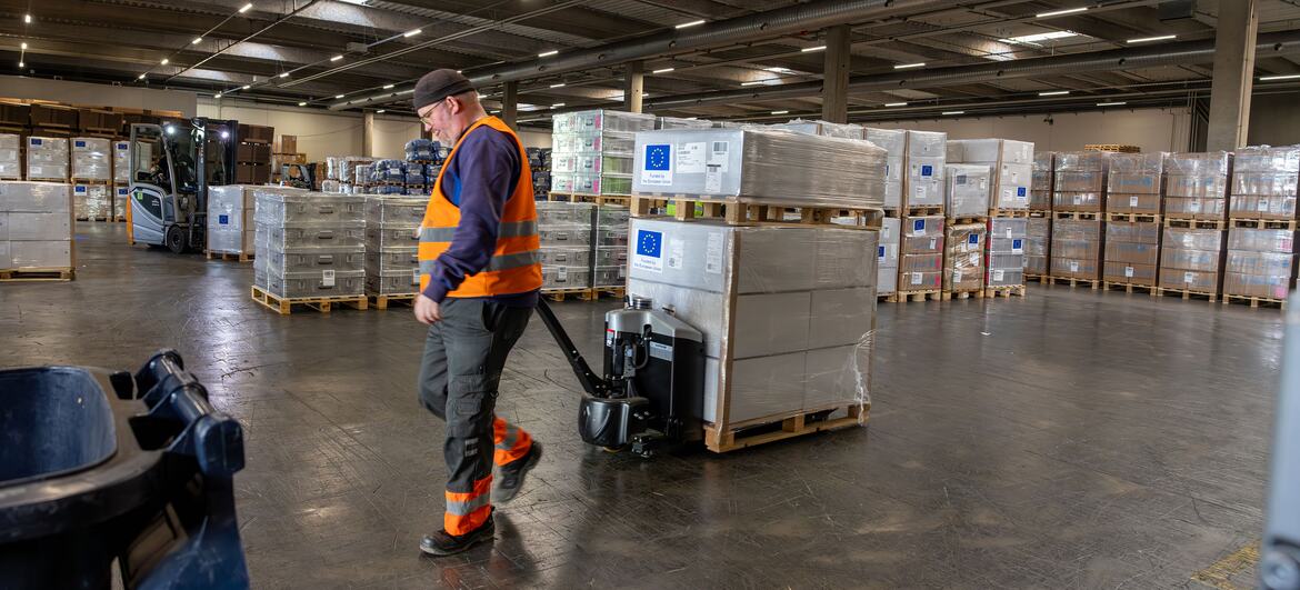 A warehouse worker in an orange safety vest pulls a pallet jack loaded with emergency supplies wrapped in plastic. The boxes feature the European Union flag. The background shows a large warehouse filled with stacks of supplies and a forklift.