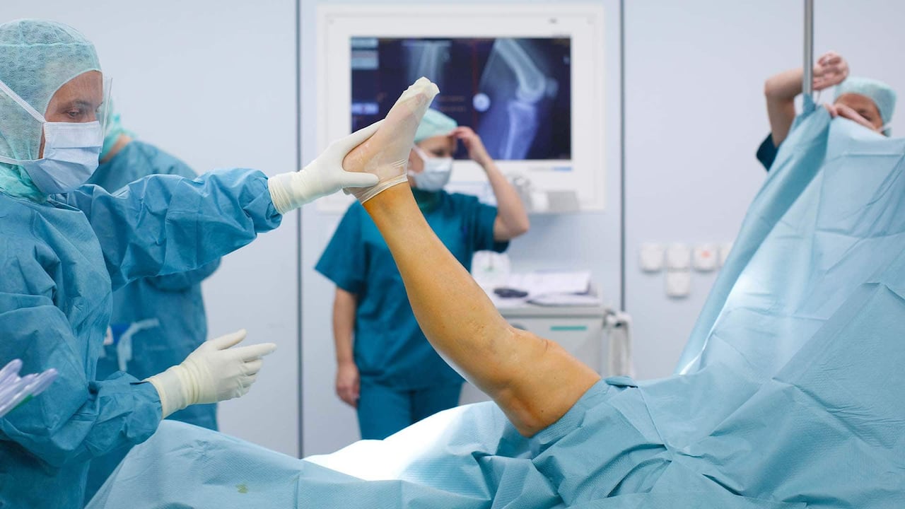 A gowned doctor holds up a patient's leg by the foot in an operating room.