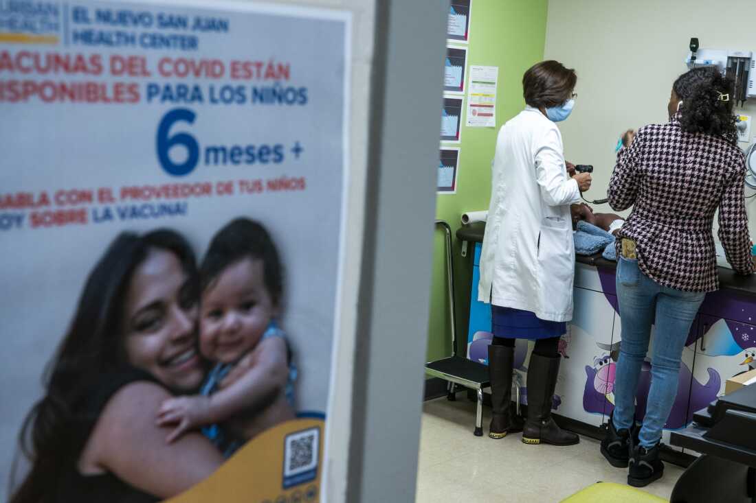 A patient care room is photographed from outside the room. A poster with Spanish language is visible, as is a doctor in a white coat, caring for a child on an exam table.