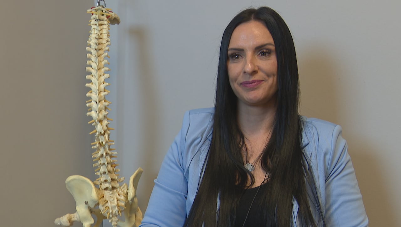 A woman with long black hair, wearing a blue suit, sits in a medical office next to a model of a spinal column and a pelvis.