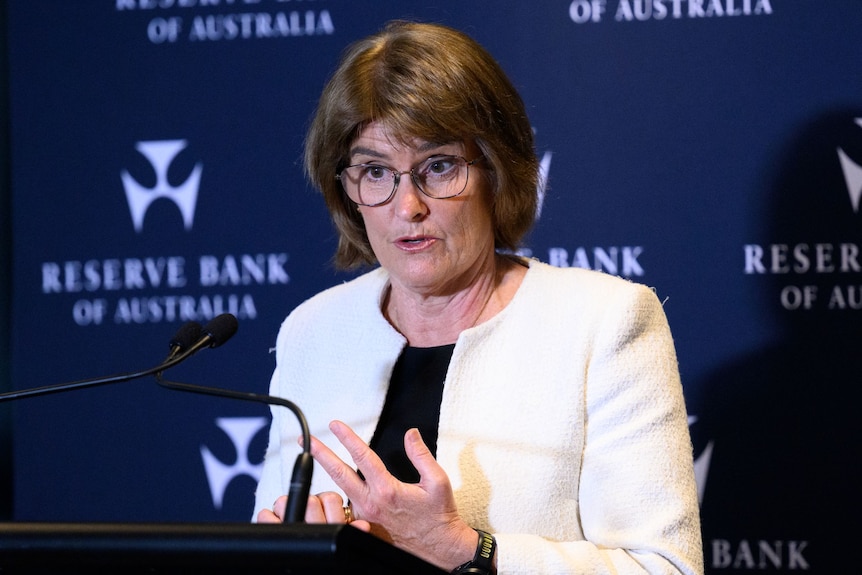 A bespectacled, auburn-haired woman in a light jacket smiles gestures as she speaks at a podium.