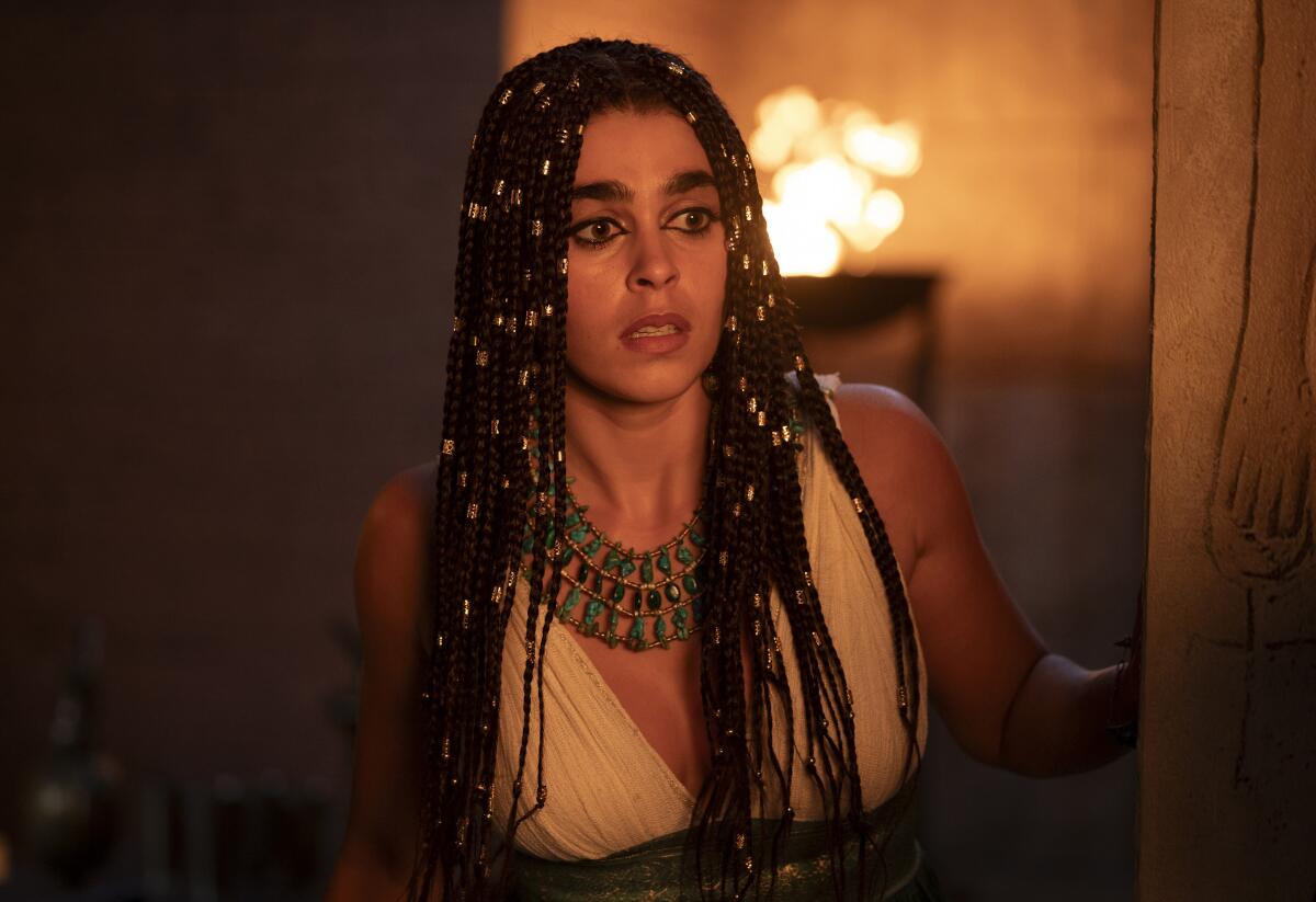 A woman with long braided dark hair, wearing a blue beaded necklace and white dress.