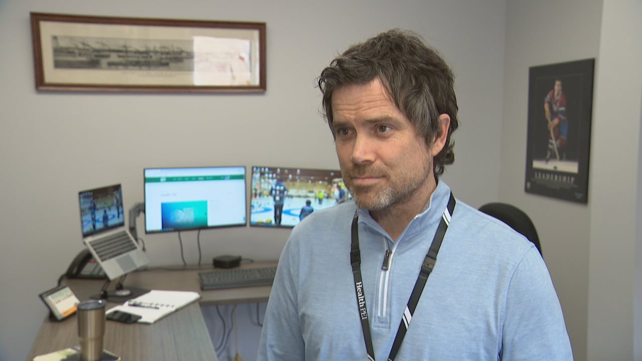 A man in a light blue shirt stands in front of a desk with a keyboard, two monitors, a laptop and a coffee mug.